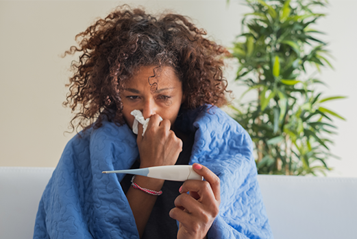 a sick women wrapped in a blanket with a tissue looking at a thermometer